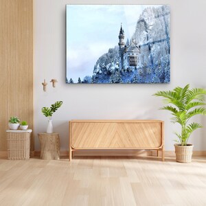 Op de afbeelding: Een ingelijste print van een besneeuwde berg met een kasteel. Het kasteel heeft een hoge toren en is omgeven door met sneeuw bedekte bomen. De print wordt weergegeven boven een houten kast in een kamer met houten vloeren.