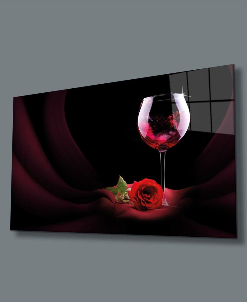 May include: A single red rose with green leaves lies on a dark red fabric next to a glass of red wine. The wine glass is clear and has a round bowl. The background is black.