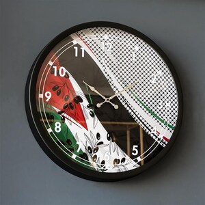 May include: A round wall clock with a black frame. The clock face features a design with red, white, black, and green elements, including olive branches and a geometric pattern. The clock hands are silver.