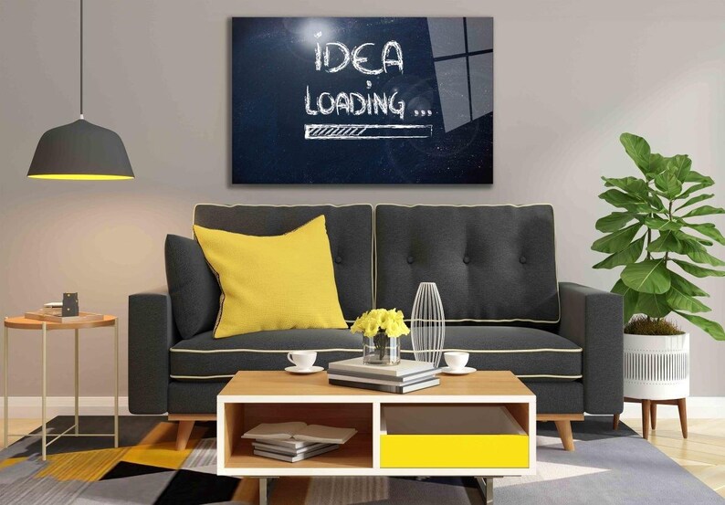 May include: A gray sofa with a yellow throw pillow and a coffee table with books and a plant. A black and white framed picture hangs on the wall above the sofa. The picture says "Idea Loading..." with a progress bar.