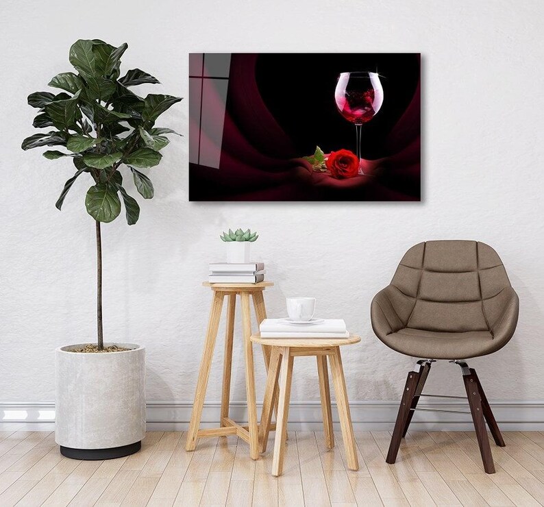 May include: A glass of red wine with a single red rose on a dark red background. The glass is on a white table with a potted plant and a chair in the background.