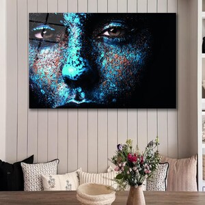May include: Close-up of a person's face with blue and brown paint splatter. The person's eyes are open and looking directly at the viewer. The background is black.