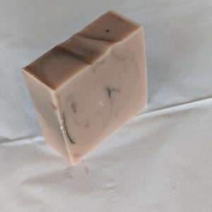 Pode incluir: Uma barra retangular de sabonete artesanal com base rosa claro e marmoreio marrom. O sabonete tem uma textura suave e um topo ligeiramente inclinado, repousando sobre uma superfície branca. O design do sabonete é simples e elegante.