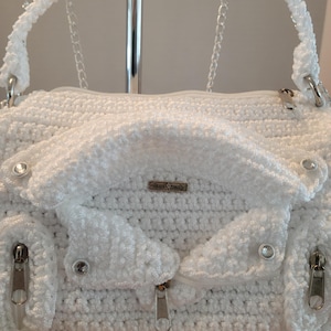 May include: A white crocheted handbag with silver-toned zippers and hardware. The bag features a textured, fluffy design, a top handle, and a chain strap. The label reads "Hand Made".