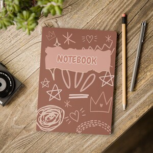 May include: A brown notebook with a light pink torn paper graphic in the center featuring the word NOTEBOOK in white letters. The cover is decorated with white line art drawings of stars, hearts, crowns, and abstract shapes, accompanied by a pencil.