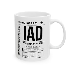 May include: A white ceramic mug featuring a black printed boarding pass design for IAD Washington DC, United States. The graphic includes a barcode, an aeroplane icon, coordinates 38° 56' 56" N, 77° 26' 57" W, and boxes for Flight VS22, Gate A14, and Seat 1A.