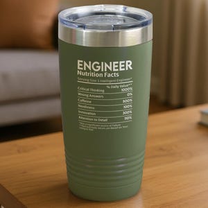 Op de afbeelding: Olijfgroene geïsoleerde beker met een zilveren rand en deksel. De beker heeft de tekst "ENGINEER Nutrition Facts" met een humoristische lijst van "dagelijkse waarden". De beker staat op een houten tafel. Hoogte: ongeveer 18 cm.