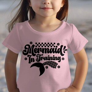 Puede incluir: Camiseta rosa claro con la frase "Mermaid In Training" en negro con una fuente estilizada. El diseño incluye una cola de sirena y elementos decorativos. La camiseta es de cuello redondo.