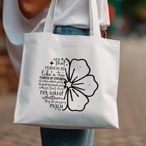 Op de afbeelding: Witte draagtas met een zwart bloemmotief en de tekst "Die persoon is als een boom geplant bij waterstromen, die zijn vrucht geeft op zijn tijd, en waarvan het blad niet verwelkt; al wat hij doet, voorspoedig is. Psalm 13."