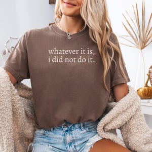 May include: A brown short-sleeved t-shirt featuring the white text "whatever it is, i did not do it." printed on the chest. The garment has a crew neckline and a relaxed fit, worn with light blue denim shorts and a textured beige cardigan.
