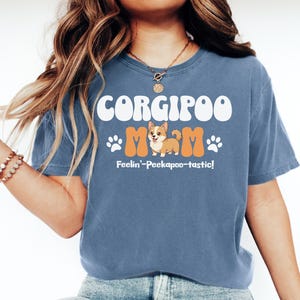 May include: Blue t-shirt with the text "CORGIPOO MOM" in white and orange. The shirt features a cartoon corgi dog and paw prints. The phrase "Feelin'-Peekapoo-tastic!" is printed below the graphic.