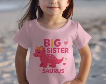Op de afbeelding: Een lichtroze T-shirt met korte mouwen en een afbeelding van een roze dinosaurus met een gele strik. De tekst BIG SISTER SAURUS staat in dikke roze letters op de voorkant, versierd met kleine roze hartjes en gele sterretjes.