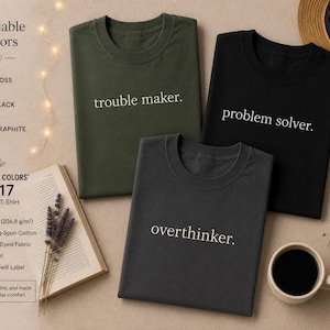 May include: Three Comfort Colors 1717 unisex t-shirts in moss, black, and graphite, each with a different embroidered word: "trouble maker," "problem solver," and "overthinker." The shirts are displayed with a book, coffee cup, and a straw hat.