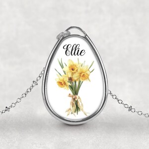 Op de afbeelding: Zilverkleurige hangerketting met een ovaalvormige bedel. De bedel toont een aquarel illustratie van gele narcissen vastgebonden met een lint, met de naam "Ellie" in zwart schrift. De ketting is delicaat.