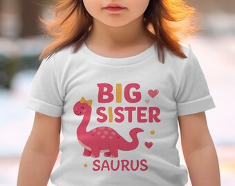 Op de afbeelding: Wit T-shirt met de woorden "BIG SISTER SAURUS" in roze en geel, met een roze dinosaurus illustratie. Het ontwerp bevat harten en sterren. Het shirt heeft korte mouwen.