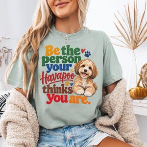 May include: A sage green t-shirt featuring a graphic of a brown Havapoo dog and the multi-coloured text: Be the person your Havapoo thinks you are. The design includes a small red heart, a blue paw print, and a yellow paw print accent.