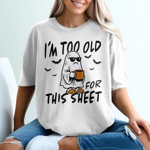 May include: White t-shirt with the phrase "I'm too old for this sheet" in black text. The graphic features a ghost wearing sunglasses and holding a mug, with bats around it. The shirt is a casual, comfortable style.