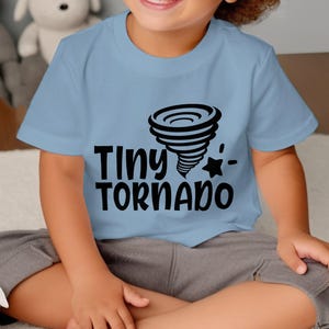 Puede incluir: Una camiseta azul claro con las palabras "Tiny Tornado" en negro, un gráfico de tornado negro y una estrella. La camiseta la lleva un niño.