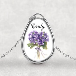 以下が含まれることがあります： 銀色のペンダントネックレス。涙型のチャームには、紫色の花の水彩画と「Emily」という名前が黒い文字で描かれています。チェーンは繊細で、背景は柔らかな白です。