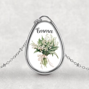 May include: Silver-toned pendant necklace with a teardrop-shaped charm. The charm features a watercolor illustration of a lily of the valley bouquet tied with a ribbon. The name "Emma" is written in script above the flowers.