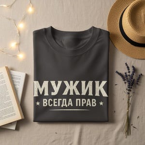 May include: Dark grey t-shirt with the Russian text "МУЖИК ВСЕГДА ПРАВ" in white, meaning "A Man is Always Right". The shirt is folded on a beige surface, with a straw hat, a book, and lavender sprigs nearby.