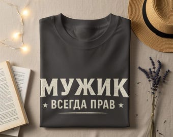 May include: Dark gray t-shirt with the Russian text "МУЖИК ВСЕГДА ПРАВ" in white, meaning "A Man is Always Right". The shirt is folded on a beige surface, with a straw hat, a book, and lavender sprigs nearby.
