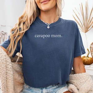 Op de afbeelding: Een marineblauw T-shirt met korte mouwen en de tekst "cavapoo mom." in het wit op de borst gedrukt. Het shirt heeft een ronde hals en een relaxte pasvorm, gemaakt van een effen gekleurde stof met een subtiele, licht versleten textuur.