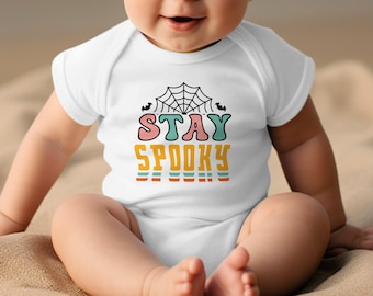 May include: White baby bodysuit with the words "STAY SPOOKY" in a colourful retro font. The design includes a spiderweb and bats, perfect for Halloween. The bodysuit is short-sleeved and suitable for infants.