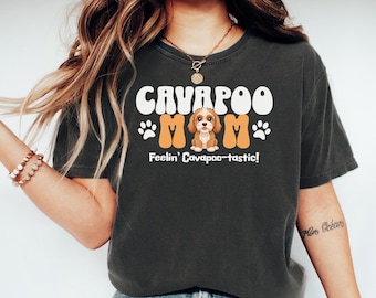 May include: Dark gray t-shirt with the words "CAVAPOO MOM" in white and orange, featuring a cartoon Cavapoo dog illustration. The text "Feelin' Cavapoo-tastic!" is below the dog graphic. The shirt is a casual style.