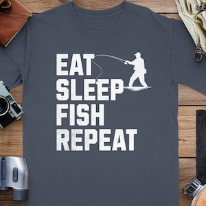 Puede incluir: Camiseta gris oscuro con las palabras "EAT SLEEP FISH REPEAT" en letras blancas. Una silueta blanca de una persona pescando está en el lado derecho del texto. La camiseta se muestra sobre una superficie de madera.