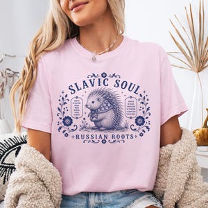 May include: A light pink t-shirt featuring a detailed illustration of a hedgehog wearing a scarf, surrounded by floral elements and the text "SLAVIC SOUL + RUSSIAN ROOTS". The design is in navy blue.