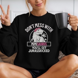 Puede incluir: Sudadera negra con el texto "DON'T MESS WITH MIMISAURUS YOU'LL GET JURASSKICKED". El gráfico presenta un dinosaurio de dibujos animados con un pañuelo. La persona sostiene una taza negra.