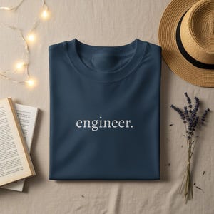 May include: A navy blue t-shirt with the word "engineer" printed in white. The shirt is folded neatly on a beige surface, with a straw hat and a sprig of lavender nearby. A book and string lights are also in the image.