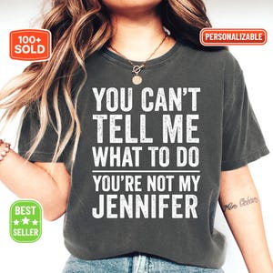 May include: Dark gray t-shirt with the text "YOU CAN'T TELL ME WHAT TO DO YOU'RE NOT MY JENNIFER" in white. The shirt has a crew neck and short sleeves. The image also includes the text "100+ SOLD", "PERSONALIZABLE", and "BEST SELLER".