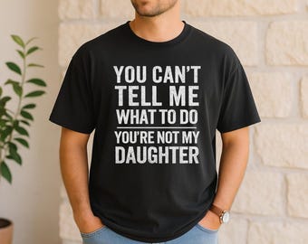 May include: Black t-shirt with the white text "YOU CAN'T TELL ME WHAT TO DO YOU'RE NOT MY DAUGHTER". The shirt is a crew neck and has short sleeves. The person is wearing a watch with a brown leather band.