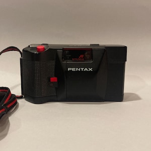 Op de afbeelding: Een zwarte Pentax-camera met een rode knop en band. De camera heeft een ingebouwde flitser en een autofocuslens.