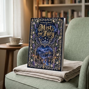 Könnte beinhalten: Ein rechteckiges Taschenbuch-Malbuch mit dem Titel A Colouring Book of Mist and Fury. Das dunkelblaue Cover zeigt eine komplexe Illustration einer Fantasiestadt, Bergen und Flügeln, eingerahmt von floralen Ranken und goldenen Akzenten.