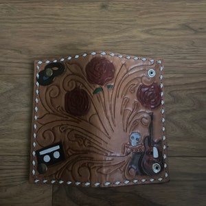 May include: A brown leather wallet with intricate floral tooling, featuring a skull, guitar, and other decorative elements. The wallet has white stitching around the edges.