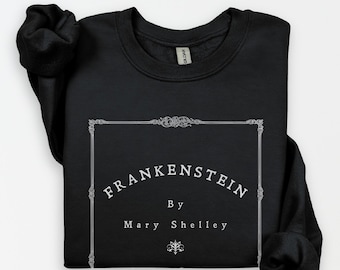 May include: Black sweatshirt featuring the text "FRANKENSTEIN by Mary Shelley" in a decorative frame. The text is in white, contrasting against the dark fabric. The sweatshirt has a crew neck and long sleeves.
