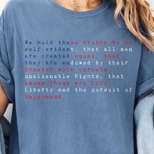 May include: A blue jean colored short-sleeve t-shirt featuring white and red text that reads: We hold these truths to be self-evident, that all men are created equal, that they are endowed by their Creator with certain unalienable Rights, that among these are Life, Liberty and the pursuit of Happiness.