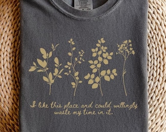 May include: A dark gray t-shirt with a crew neck, featuring a gold botanical design and the quote, "I like this place and could willingly waste my time in it." The shirt is folded on a woven surface.