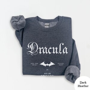 Op de afbeelding: Donkergrijs sweatshirt met het woord "Dracula" in gotische letters. Daaronder staat een vleermuisafbeelding en de tekst "FOR THE DEAD TRAVEL FAST BY BRAM STOKER". De sweatshirt heeft een ronde hals en opgevouwen mouwen.