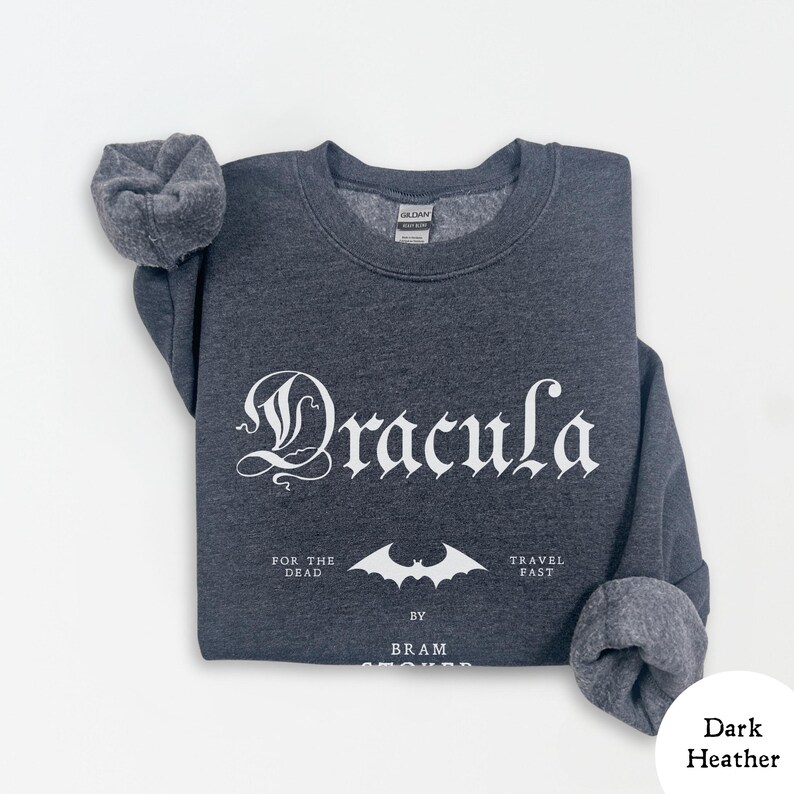 Op de afbeelding: Donkergrijs sweatshirt met het woord "Dracula" in gotische letters. Daaronder staat een vleermuisafbeelding en de tekst "FOR THE DEAD TRAVEL FAST BY BRAM STOKER". De sweatshirt heeft een ronde hals en opgevouwen mouwen.