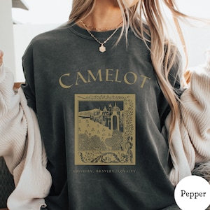 May include: Dark gray t-shirt with the word "CAMELOT" in gold lettering. Below the word is a gold-colored illustration of a castle and trees, with the words "CHIVALRY, BRAVERY, LOYALTY" at the bottom.