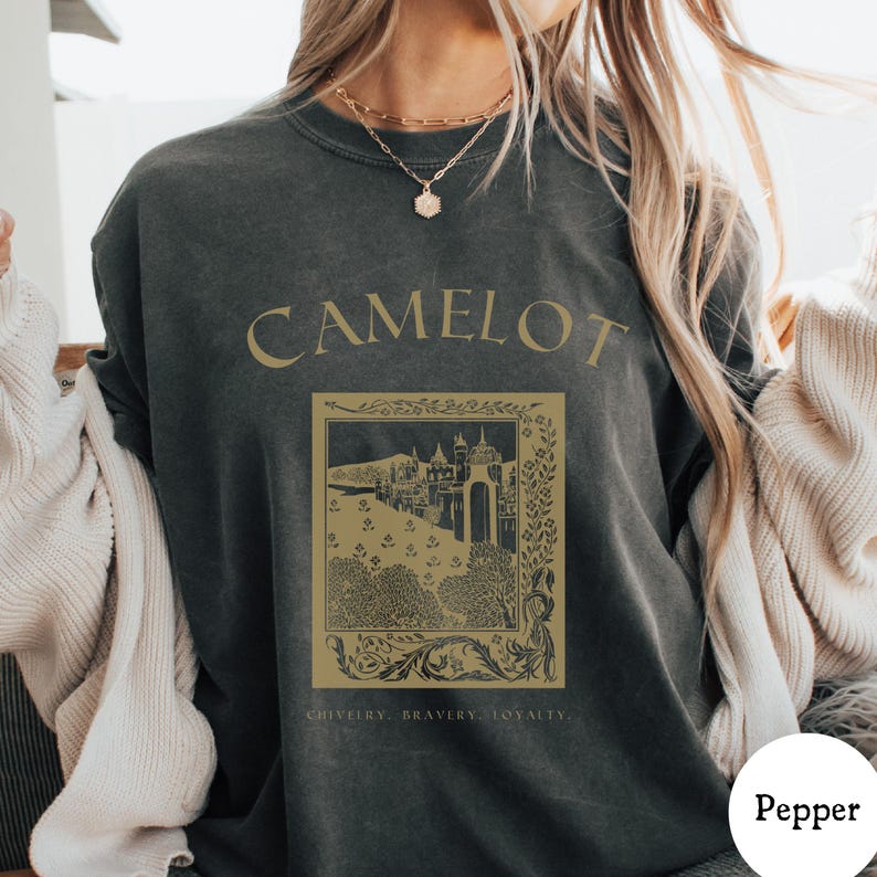 May include: Dark gray t-shirt with the word "CAMELOT" in gold lettering. Below the word is a gold-colored illustration of a castle and trees, with the words "CHIVALRY, BRAVERY, LOYALTY" at the bottom.