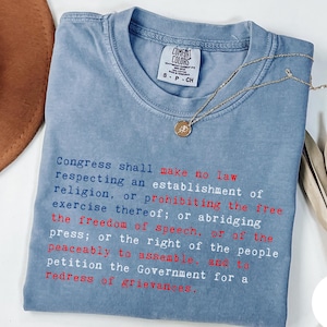 May include: A blue Comfort Colors t-shirt with the First Amendment text printed in red and blue. A gold necklace with a pendant is draped across the shirt. The shirt is folded on a white surface.