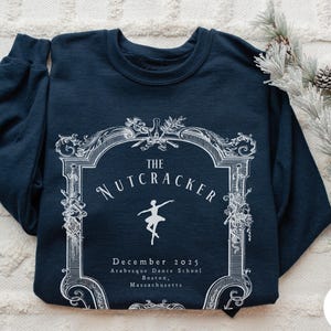 May include: A navy blue crewneck sweatshirt featuring a white graphic print of an ornate frame. Inside the frame is the text "THE NUTCRACKER", a silhouette of a ballet dancer, and the text "December 2025 Arabesque Dance School Boston, Massachusetts".