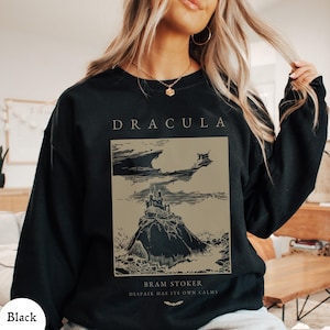 May include: A black long-sleeve sweatshirt featuring a tan rectangular graphic print. The text reads DRACULA at the top and BRAM STOKER DESPAIR HAS ITS OWN CALMS at the bottom. The graphic depicts a castle on a hill under a dark, cloudy sky.