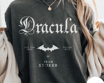 May include: A charcoal gray crewneck t-shirt featuring the white text Dracula, For The Dead, Travel Fast, and Bram Stoker, along with a central white bat graphic. The shirt is layered under a beige cardigan and paired with a gold chain necklace.