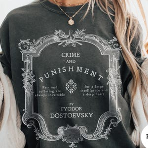 May include: A charcoal gray crewneck sweatshirt featuring a white graphic print of an ornate frame containing the text Crime and Punishment, Pain and suffering are always inevitable for a large intelligence and a deep heart, by Fyodor Dostoevsky.